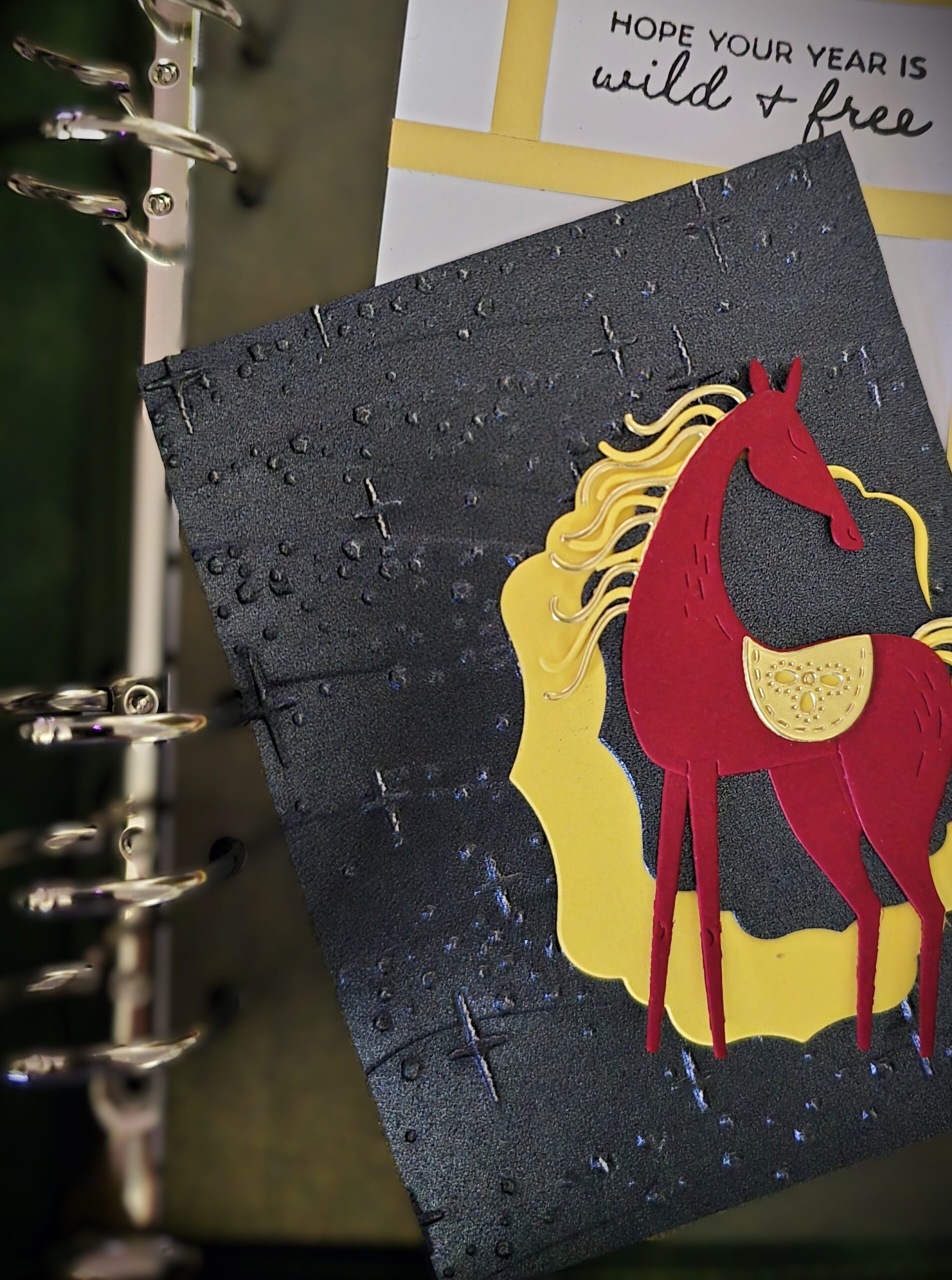 Handcrafted journal page featuring a layered red horse with gold accents on a dark textured background, resting inside an open binder.
