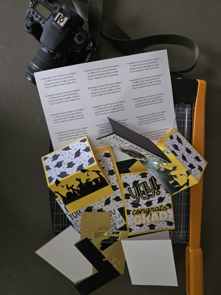 A handmade graduation card workspace with black, gold, and white cards in progress beside paper scraps and crafting tools. A printed sheet with the message “Graduation is more than just a degree…” sits beneath the cards, with a Canon camera resting nearby in soft natural light.