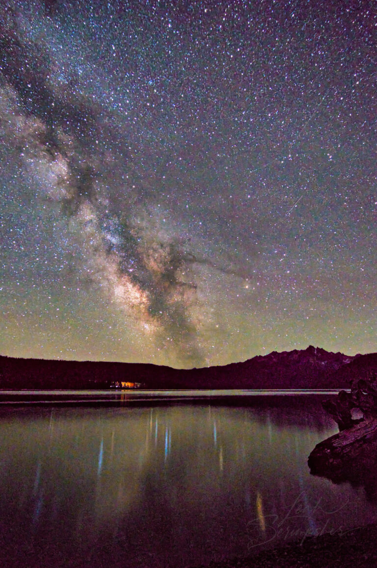 Milky Way stretching above Redfish Lake and reflected in calm water beneath Grand Mogul Peak, with a faint green aurora glow along the horizon.