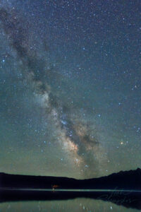 Wide view of the Milky Way above Redfish Lake, with a faint green aurora glow near the horizon and reflections shimmering on the lake beneath Grand Mogul Peak.