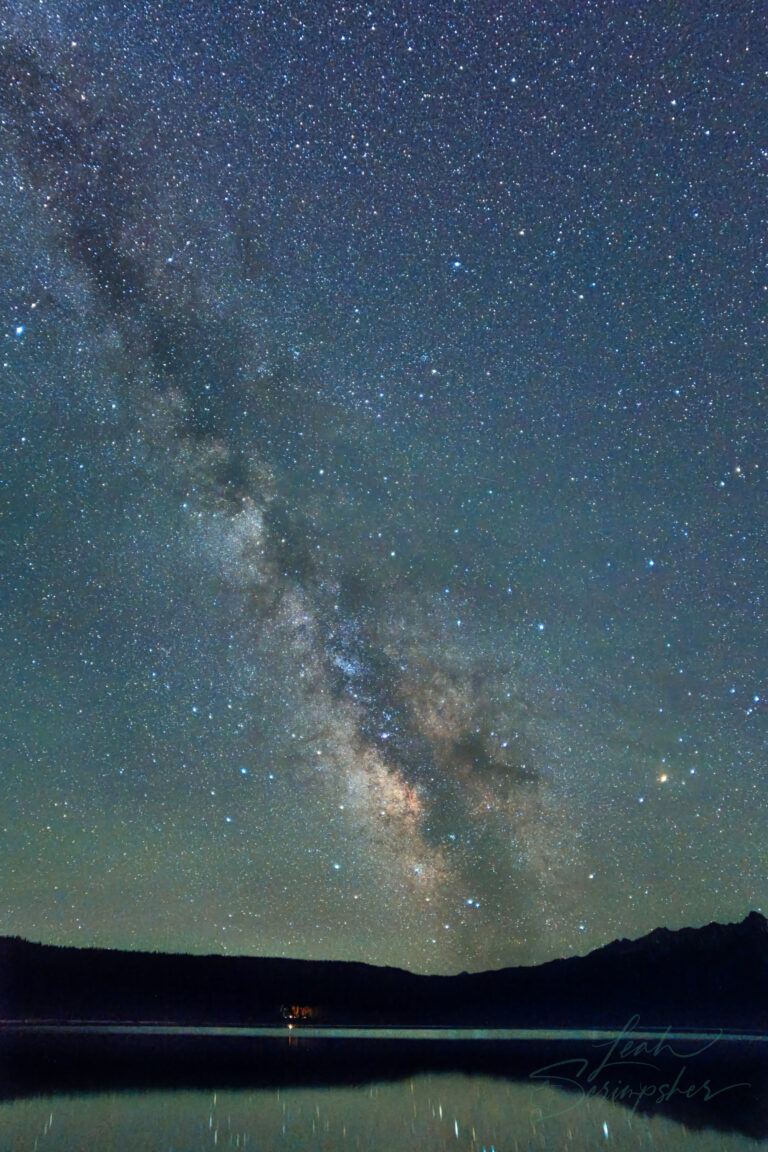 Wide view of the Milky Way above Redfish Lake, with a faint green aurora glow near the horizon and reflections shimmering on the lake beneath Grand Mogul Peak.