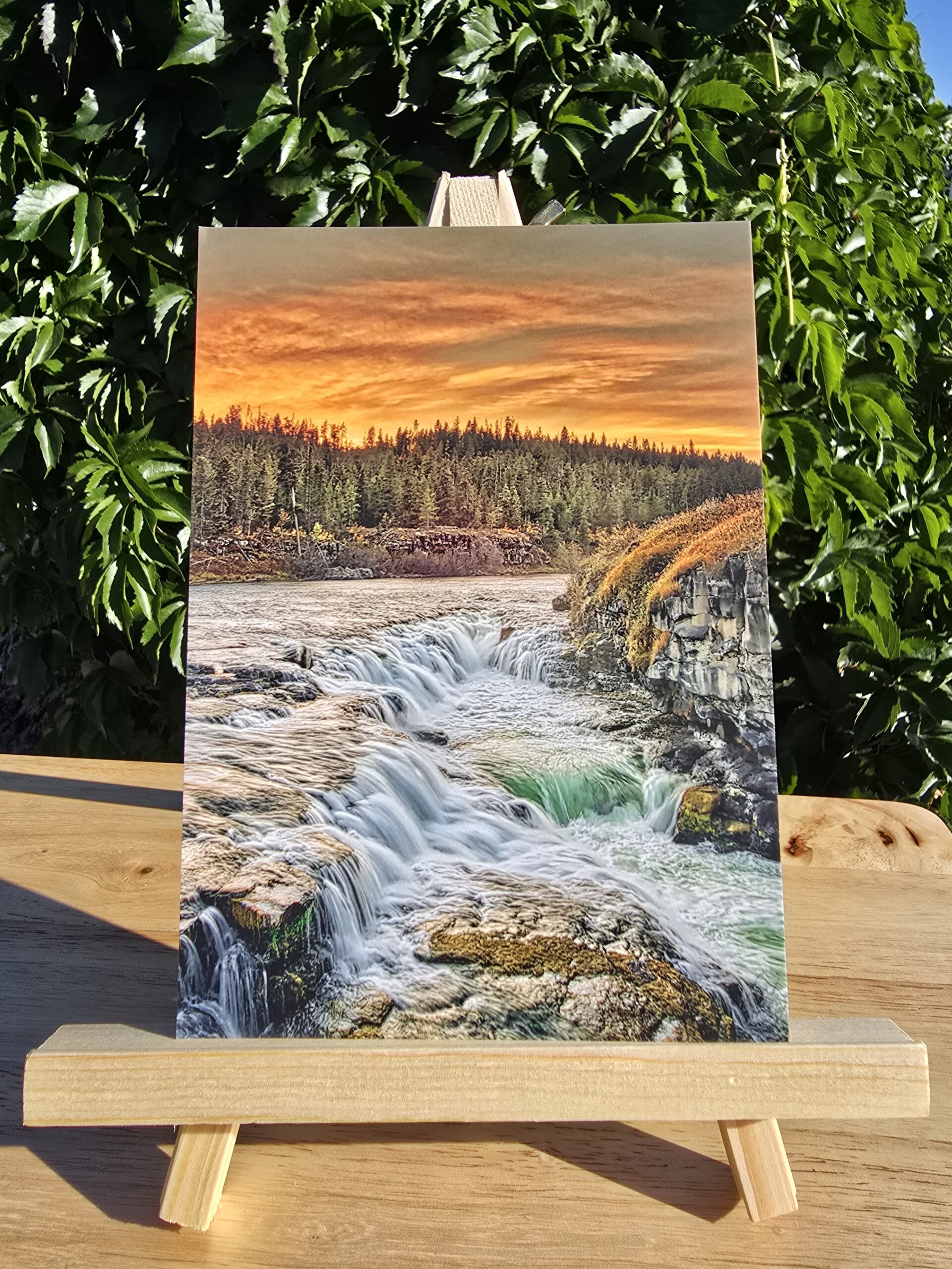 Postcard featuring Sheep Falls Sunset displayed on a small wooden easel, colorful sunset over a waterfall